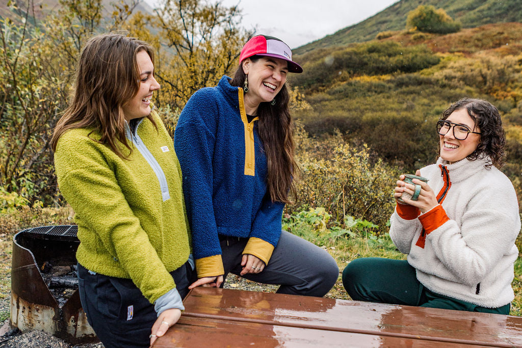Three women laughing and talking outdoors at a wooden picnic table, with a metal grill on the left and a grassy hill in the background. They are wearing cozy fleece jackets in lime green, blue with yellow trim, and cream with orange trim.