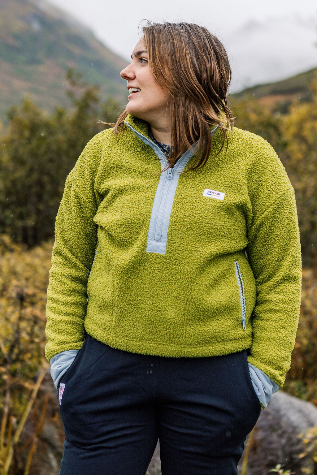 A woman wearing a lime green fleece pullover with a light blue zipper placket, standing outdoors in a mountainous, forested environment.