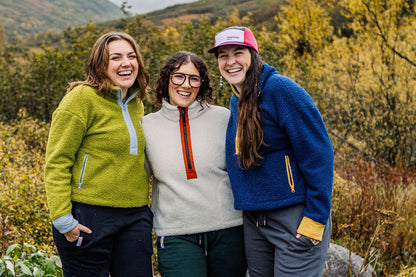 Three women smiling outdoors in autumn foliage, wearing fleece pullovers in green, cream with an orange zipper, and blue, with one wearing a pink and white cap.