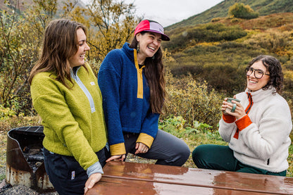 Three women laughing and talking outdoors at a wooden picnic table, with a metal grill on the left and a grassy hill in the background. They are wearing cozy fleece jackets in lime green, blue with yellow trim, and cream with orange trim.