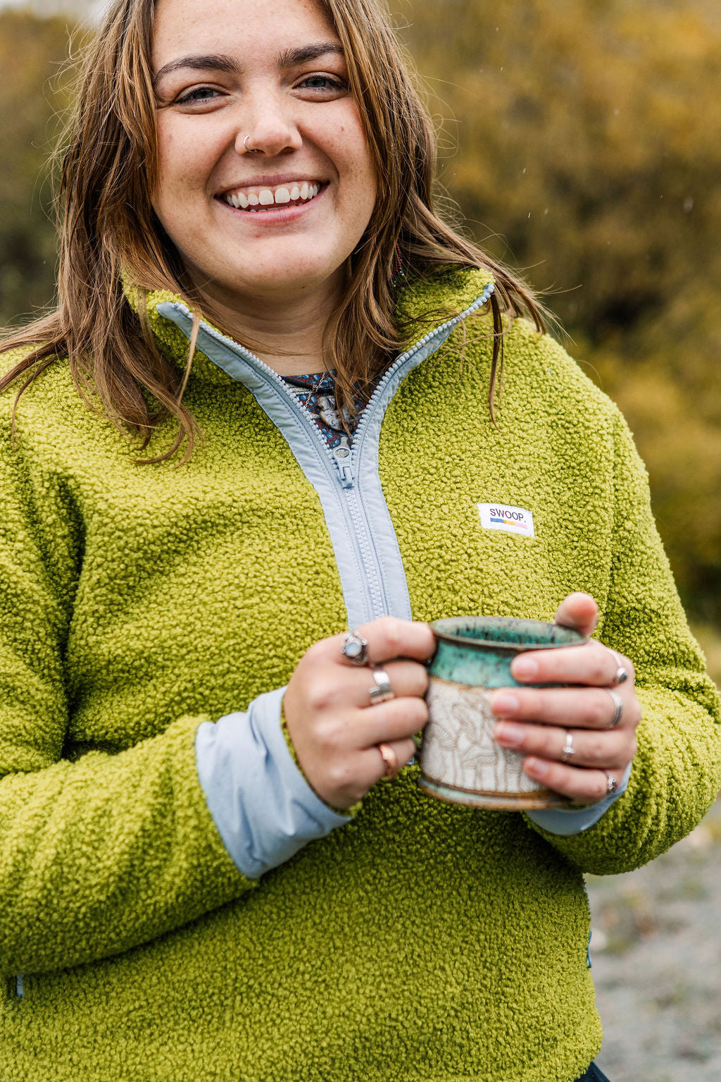 A smiling woman outdoors wearing a lime green fleece zip-up jacket with a light blue zipper and holding a ceramic mug.