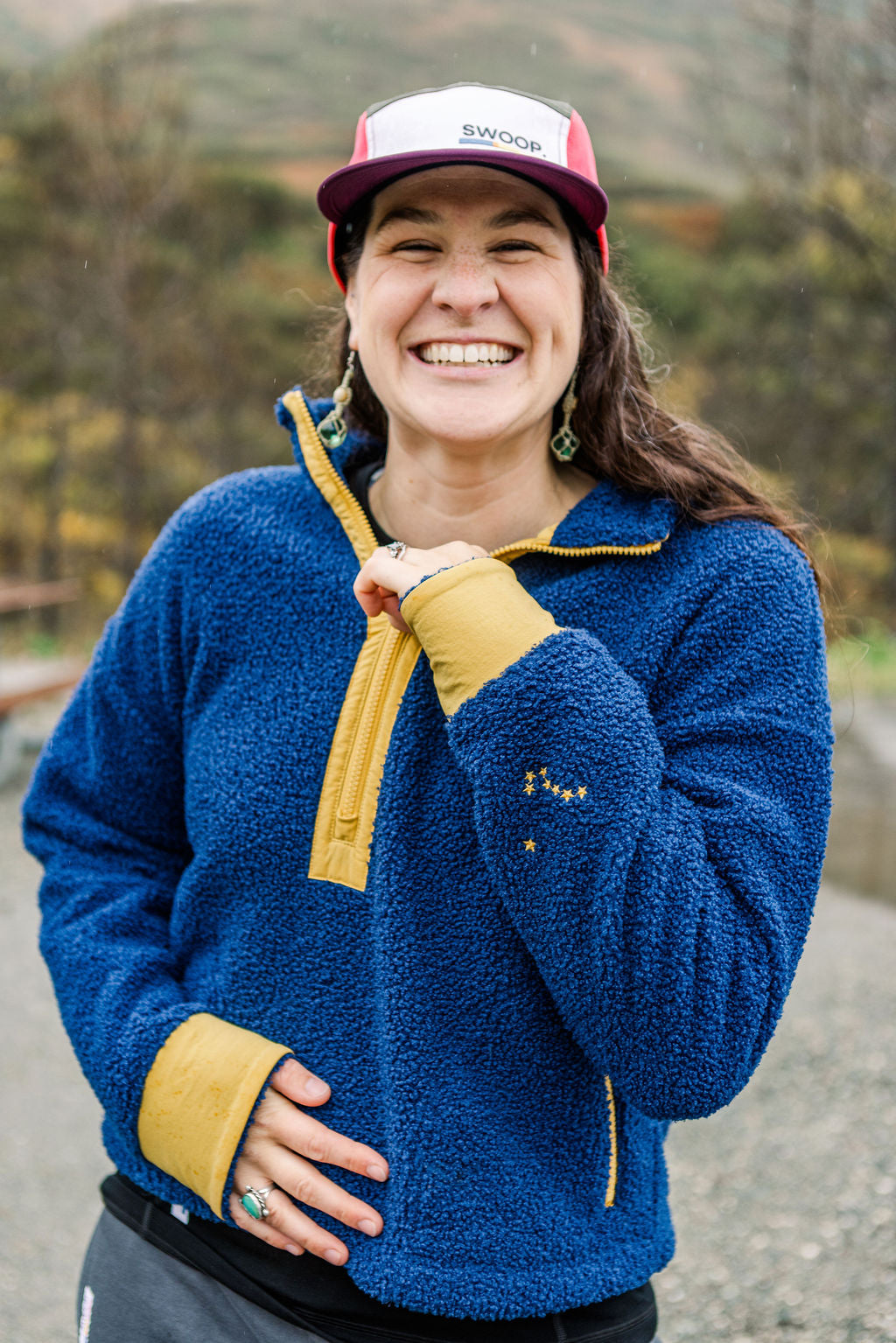 A smiling woman outdoors wearing a blue fleece pullover with mustard yellow trim and matching cuffs, along with a red and white trucker hat. The background shows a blurred natural setting with hills and trees.
